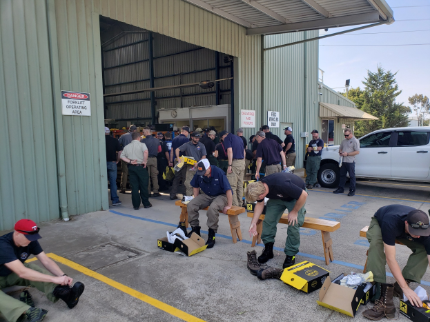 Eine Gruppe von Menschen bei einer Ausbildungssitzung für die Nationalgarde, einige sitzen auf Bänken und andere stehen, mit verstreuten Kartonagen, Schuhen und Gegenständen vor einem Gebäude, Fahrzeugen und Bäumen im Hintergrund unter einem klaren blauen Himmel.