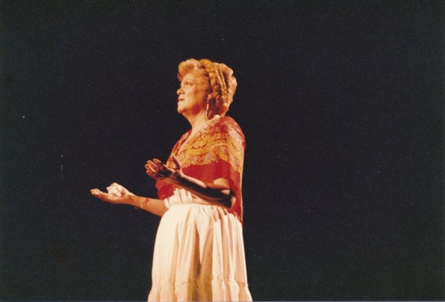 Eine Frau in einem weißen, hochgeschlossenen, langärmeligen Kleid steht auf einer Bühne mit ausgebreiteten Armen, beleuchtet gegen einen dunklen Hintergrund, ihre Haare zu einem Dutt gebunden und einen ernsten Gesichtsausdruck.