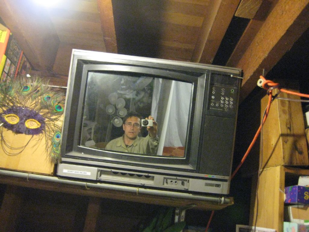 Ein Fernsehbildschirm zeigt eine Person umgeben von Pfauenfedern, Büchern und Kisten.