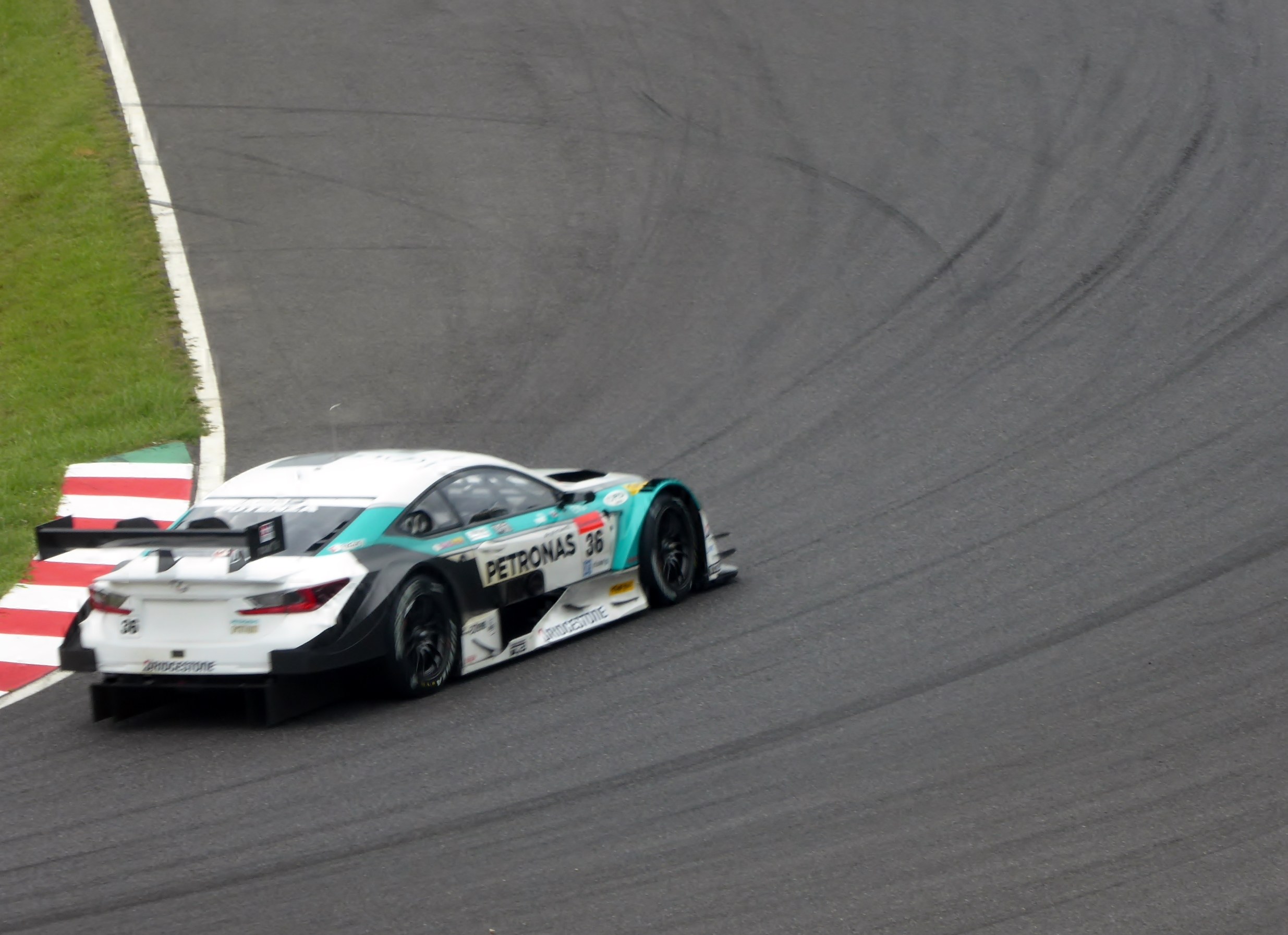 Ein silberner Mercedes-AMG GT3-Rennwagen mit Sponsorentext auf der Karosserie, der auf einer Rennstrecke fährt, mit Gras links sichtbar.