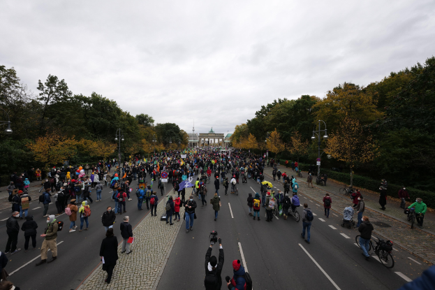 Eine große Gruppe von Menschen marschiert eine baumbestandene Straße in Berlin entlang, einige halten Kameras, mit einem Gebäude und einem klaren Himmel im Hintergrund.