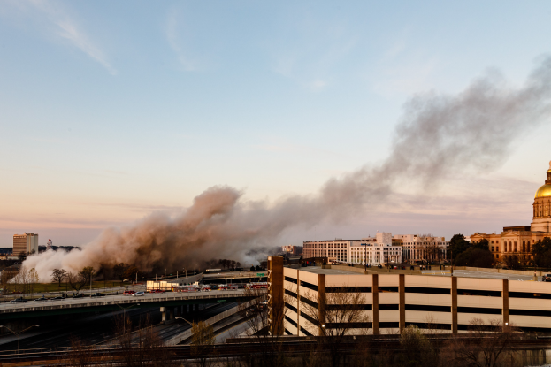 Eine dicke Rauchwolke steigt aus einem Gebäude in einer Stadtlandschaft auf, umgeben von Bäumen, Fahrzeugen, Straßenlaternen und einer Brücke, mit dem Himmel im Hintergrund.
