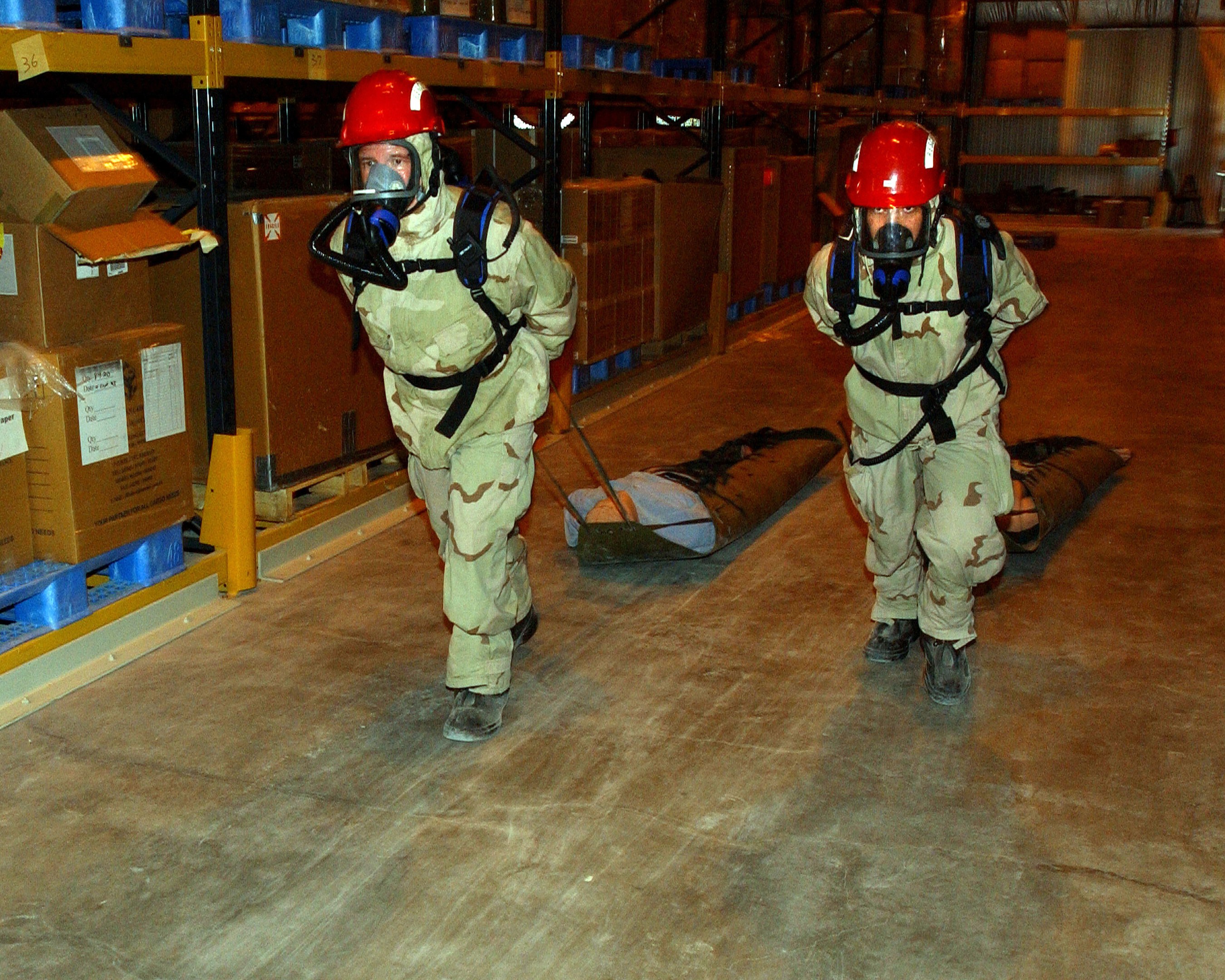 Zwei Militärangehörige in roten Helmen und Gasmasken tragen eine Trage durch ein Lagerhaus voller Kartonagen und Regale.