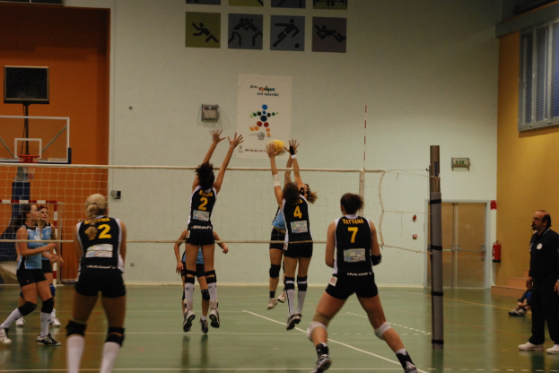 Ein Volleyballspiel auf einem Indoor-Court mit Spielern, einem Basketballkorb und einer Tür und Fenstern im Hintergrund.