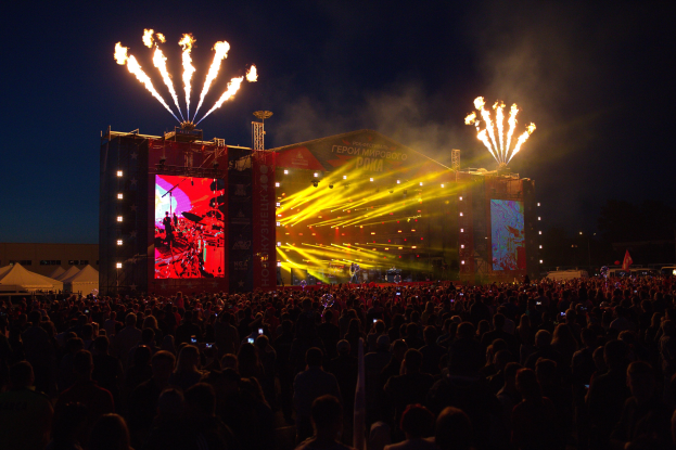 Eine große Menge bei einem Musikfestival schaut Performern auf einer hell erleuchteten Bühne zu, mit Zelten links und Feuerwerk in einem dunklen Himmel dahinter.