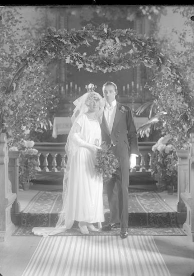 Schwarz-weißes Foto eines Mannes und einer Frau, die unter einem Blumenschmuckbogen stehen, die Frau hält einen Strauß Blumen, mit einem Teppichboden.