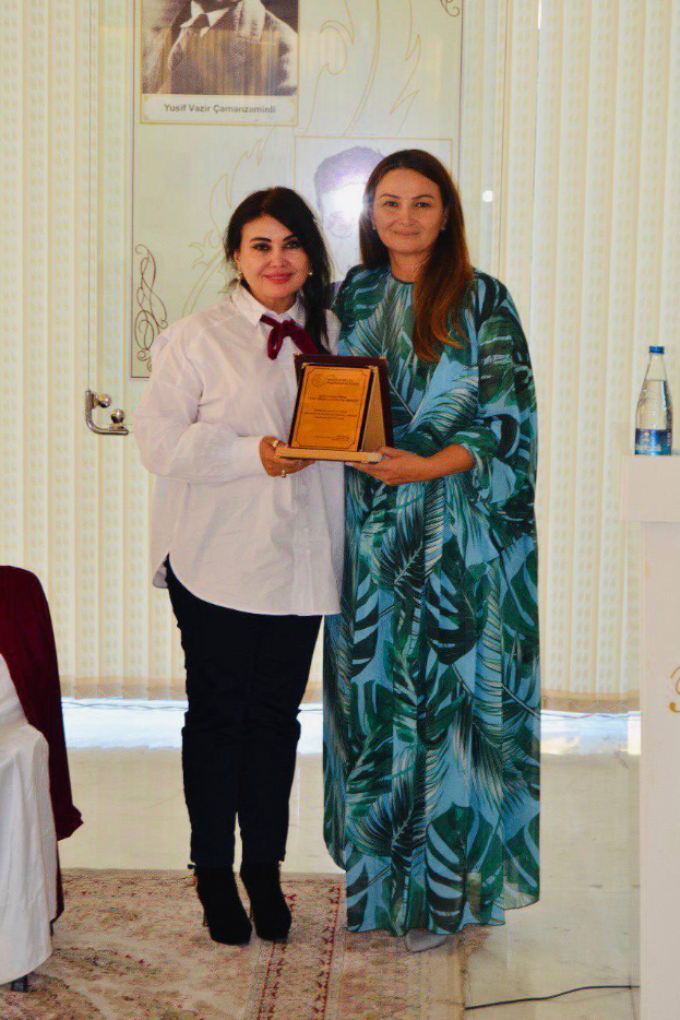 Zwei Frauen bei einer Preisverleihung, eine hält einen Preis und lächelt, die andere steht neben ihr, mit einem Rednerpult, einer Wasserflasche, Vorhängen und einem Fotorahmen im Hintergrund.