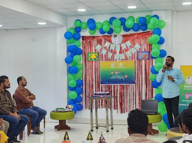 Eine Gruppe von Menschen sitzt auf Stühlen vor einer Bühne mit Ballons, einem Banner, einer Uhr und Lichtern, wo eine Person ein Mikrofon hält und ein Tisch einen Laptop und andere Gegenstände während einer 9. Geburtstagsfeier zeigt.