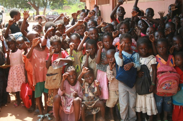 Eine Gruppe von Kindern mit erhobenen Händen, mit einem Gebäude auf der rechten Seite und einem Fahrzeug unter Bäumen im Hintergrund.