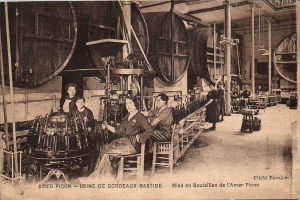 Ein Schwarz-Weiß-Foto von Frauen, die in einer Fabrik arbeiten, mit einigen, die auf Stühlen sitzen und anderen, die zwischen verschiedenen Maschinen stehen, und Text unten, der "Amer Picon - Use de Bordeaux-Bastide" lautet.