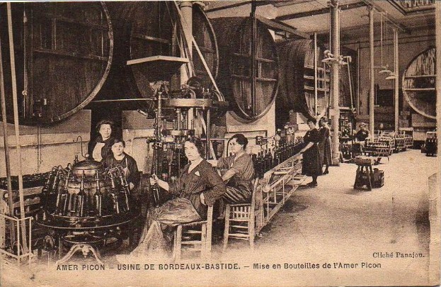 Ein Schwarz-Weiß-Foto von Frauen, die in einer Fabrik arbeiten, mit einigen, die auf Stühlen sitzen und anderen, die zwischen verschiedenen Maschinen stehen, und Text unten, der "Amer Picon - Use de Bordeaux-Bastide" lautet.