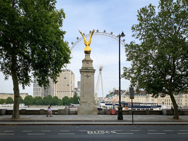 Das London Eye, ein großer Riesenrad, ist im Hintergrund zu sehen, mit einer Statue auf einem Sockel im Vordergrund, einer Straße darunter, Bäumen auf beiden Seiten, einem Laternenmast und einigen Menschen, die auf dem Gehweg spazieren gehen, mit Schiffen auf dem Wasser, mehr Bäumen, Gebäuden und dem Himmel.