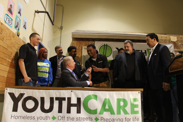 Eine Gruppe von Menschen um einen Tisch mit einer "Jugendpflege"-Aufschrift, eine Person in einem Stuhl in der Mitte, ein Podium mit einem Mikrofon davor und eine hölzerne Wand mit Papieren und Rohren auf der linken Seite.