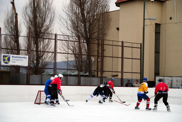 Menschen beim Eishockey auf einem Eisring mit Gebäuden, Bäumen, einer Straßenlaterne, einem Namensschild und Zäunen im Hintergrund unter einem klaren Himmel.