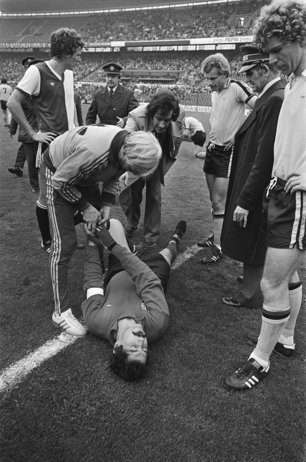 Schwarzes und weißes Foto eines Fußballspielers, der auf dem Boden liegt, während ihn eine Gruppe von Männern und ein Stadionpublikum umgibt; der Spieler scheint verletzt zu sein.