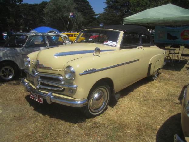 Autos stehen auf einem freien Gelände mit einem Tisch und Stühlen auf der rechten Seite, einem Zelt und Banner oben rechts und Bäumen oben im Bild.