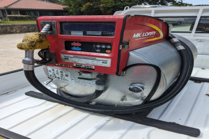 Ein Lastwagen mit einer Zapfsäule auf der Ladefläche parkt neben einem Haus, umgeben von Bäumen unter einem klaren blauen Himmel.