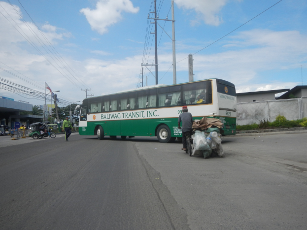 Grüner und weißer Bus fährt auf einer Straße neben einem Gebäude mit Fußgängern, einem Fahrradfahrer, Strommasten, Gebäuden, Bäumen und einer bewölkten Himmel im Hintergrund.