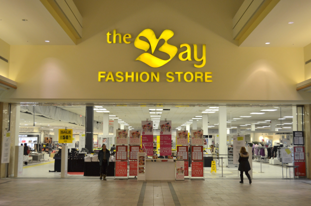 Außenansicht eines Bay Fashion Stores in einem Einkaufszentrum mit Menschen, Wandtafeln, Säulen, Deckenleuchten und Hintergrundauslagen.