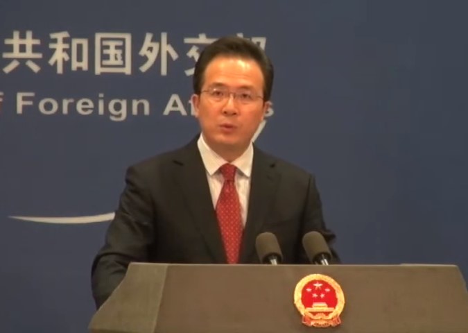 Mann im Anzug und Krawatte, der an einem Podium mit zwei Mikrofonen spricht, mit einem Logo auf dem Podium und einem Banner mit Text im Hintergrund.