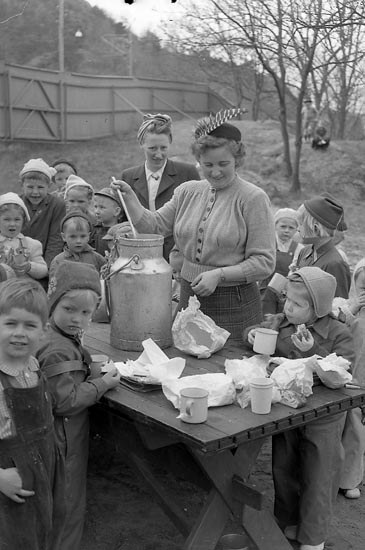 Schwarzes und weißes Bild von Kindern, die um einen Tisch versammelt sind, während eine Frau Milch aus einem Krug in einen Behälter gießt, mit Tassen und Deckeln auf dem Tisch und Bäumen im Hintergrund.