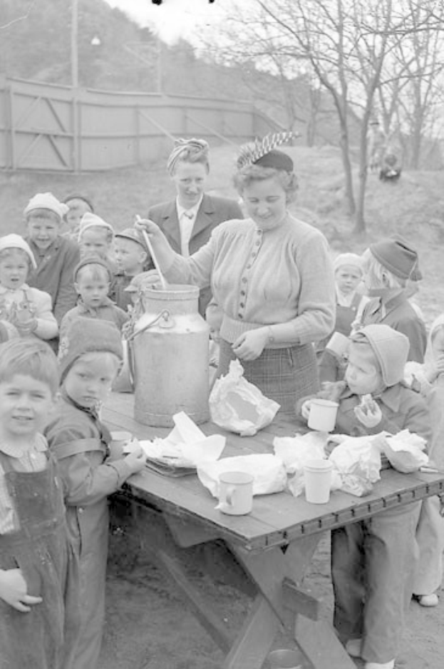 Schwarzes und weißes Bild von Kindern, die um einen Tisch versammelt sind, während eine Frau Milch aus einem Krug in einen Behälter gießt, mit Tassen und Deckeln auf dem Tisch und Bäumen im Hintergrund.