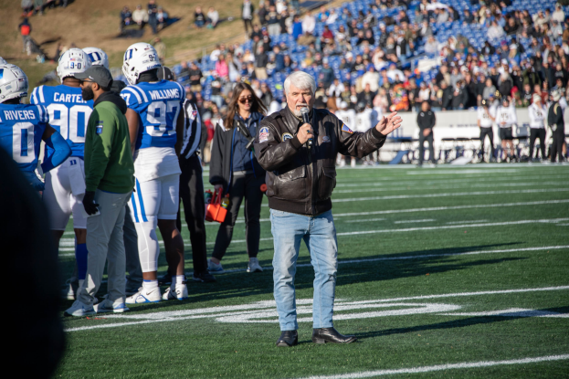 Ein Mann in schwarzer Jacke, blauen Jeans und schwarzen Schuhen steht auf einem Fussballfeld und h├Ąlt ein Mikrofon in der Hand, mit einer Menschenmenge im Hintergrund, einige tragen Helme.