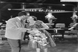 Ein Mann und eine Frau umarmen und küssen sich auf einer Bühne, wobei die Frau einen Blumenstrauß hält, während Stühle und Blumenvasen im Hintergrund sichtbar sind.