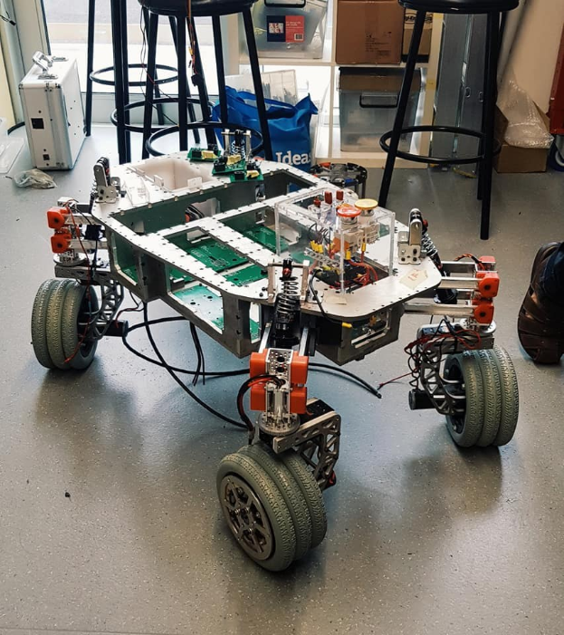 Ein Roboter sitzt auf einem Tisch in einem Raum mit Hockern, Kartons und anderen Gegenständen, mit einem Teil eines Menschenbeins auf der rechten Seite.