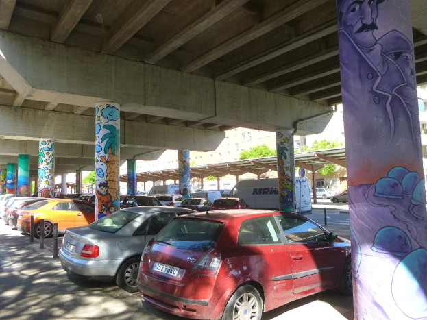 Ein Parkplatz mit zahlreichen geparkten Autos unter einer Brücke, Graffiti an den Pfeilern und Gebäuden und Bäume im Hintergrund, inklusive eines geparkten Vans.