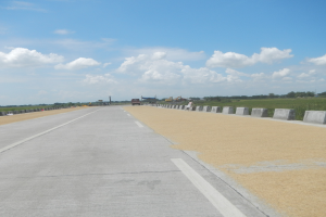 Eine Straße mit Betonleitwänden auf beiden Seiten, Fahrzeuge fahren darauf, mit Bäumen und Wolken im Hintergrund.