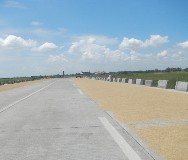 Eine Straße mit Betonleitwänden auf beiden Seiten, Fahrzeuge fahren darauf, mit Bäumen und Wolken im Hintergrund.
