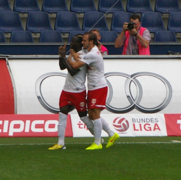 Zwei Fußballspieler feiern ein Tor auf einem Feld mit Zuschauern, einem Fotografen, einem Banner, Stühlen und einer Wand im Hintergrund.
