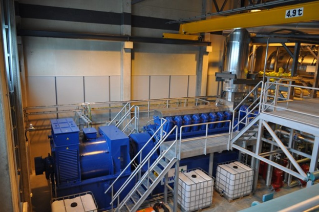 Eine Fabrikinnensicht mit Trägern, einer Treppe, einem Geländer, Containern, Lampen, Rohren und Wänden.