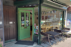 Ein Freisitz mit Tischen und Stühlen bei Green Kitchen Vegan Cafe, umgeben von Pflanzen und einer Matte, mit einer Glaswand, die Plakate zeigt und einen Blick auf den Innenbereich mit einer hängenden Lampe gewährt.