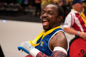 Ein Mann im Rollstuhl mit ausgestreckten Armen und einer Medaille um den Hals lächlt im Vordergrund, während im Hintergrund Menschen, Stühle und andere Gegenstände zu sehen sind.