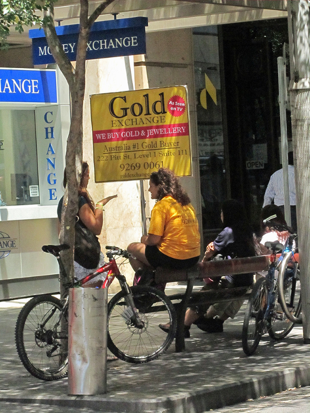 Eine Straßenszene mit Geschäften in der Mitte, einem Fahrrad, einer Bank auf der rechten Seite und einem Baum auf der linken Seite.