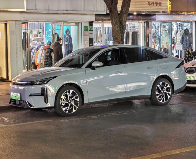 Ein Mitsubishi Lancer EV Elektroauto auf einer Stadtstraße bei Nacht geparkt, umgeben von Gebäuden, beleuchteten Schaufenstern, Schaufensterpuppen in Kleidern und Schildern.