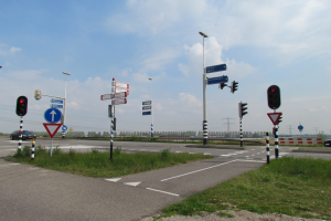 Ein Kreisverkehr mit Fahrzeugen, Gras, Ampeln, Schildern, Laternen, Strommasten mit Drähten, Bäumen und einem bewölkten Himmel; zusätzliche Ampeln befinden sich am Straßenrand.