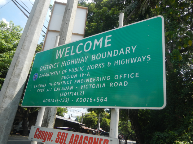 Grüne Schilder mit der Aufschrift 'Willkommen an der Bezirksgrenze' sind an zwei weißen Pfählen befestigt, mit Fahrzeugen, Gebäuden und Bäumen im Hintergrund unter einem blauen und weißen Himmel.