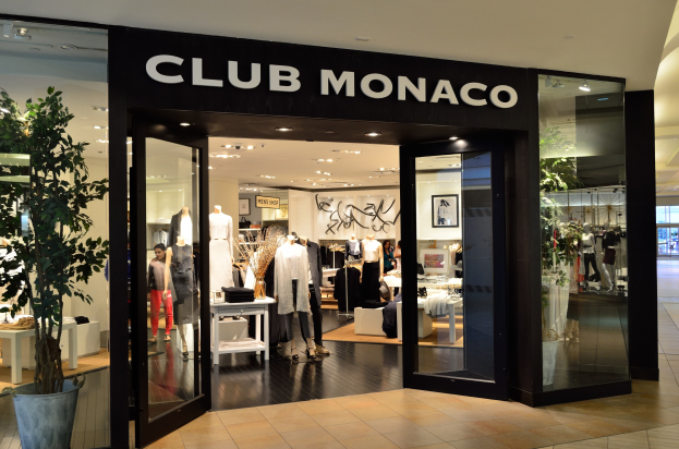 Außenansicht eines Club Monaco Geschäfts in einem Einkaufszentrum mit Glaswänden und -türen, Schaufensterpuppen in Kleidern, Auslagetischen mit Artikeln, einem Topf mit einer Pflanze und Deckenleuchten.