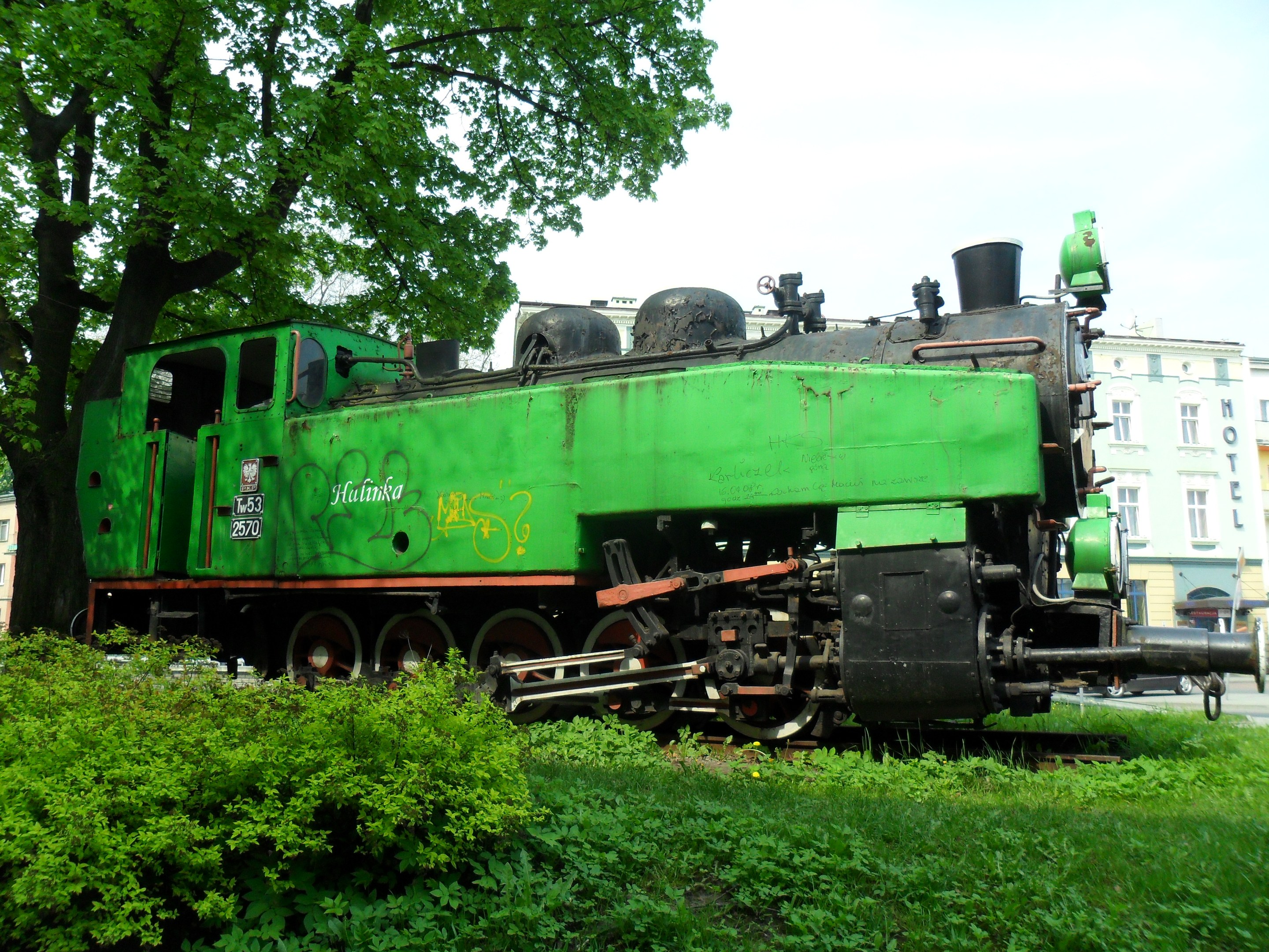 Ein grüner Zugmotor auf einem saftig grünen Feld mit Pflanzen und Bäumen, Gebäuden, Fahrzeugen und einem klaren blauen Himmel im Hintergrund.