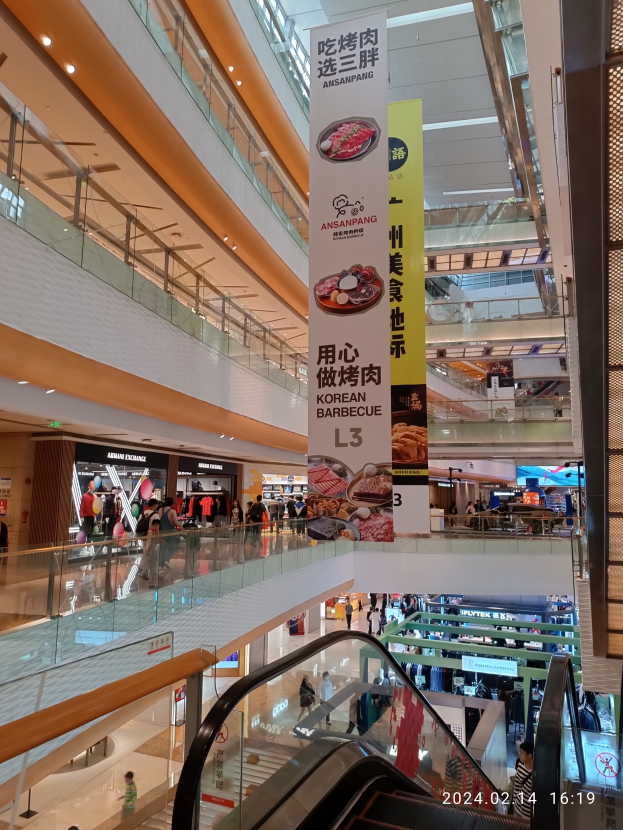 Ein Innenblick in ein Einkaufszentrum mit einer Rolltreppe in der Mitte, Menschen, die herumlaufen, einige mit Handtaschen, und Banner an der Decke hängen, mit Geschäften im Hintergrund.