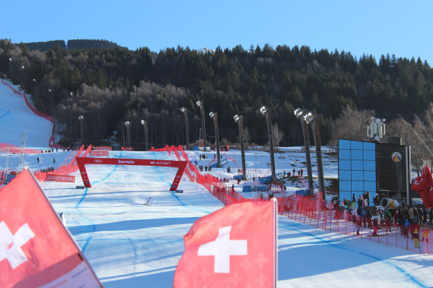 Eine Gruppe von Menschen, die auf einem schneebedeckten Hang mit roten Fahnen im Vordergrund, Laternen, Türen und einem klaren blauen Himmel im Hintergrund und einem große Bildschirm auf der rechten Seite stehen.