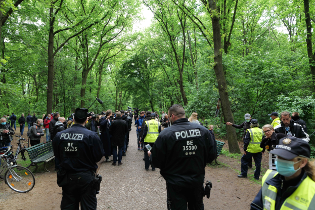 Eine Gruppe von Polizisten vor einer Menschenmenge während einer Anti-Terror-Demonstration in Berlin, mit Fahrrädern und einer Bank im Vordergrund und Bäumen und Himmel im Hintergrund.