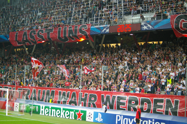 Ein Fußballspiel in einem Stadion mit einer großen Zuschauermenge, grünem Rasen, einem Torpfosten links, Bannern auf dem Boden und Menschen mit Fahnen, mit Geländern, Tafeln und Lichtern im Hintergrund.