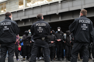 Eine Gruppe von Polizisten in Uniform steht vor einer Menge von Menschen, die ebenfalls schwarze Uniformen und Masken tragen, mit einer Brücke und einem Gebäude im Hintergrund, während einer Demonstration in einer Stadt.