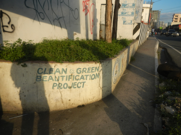 Eine Straße mit Fahrzeugen, Gebäuden, Bäumen, Polen, Drähten und einer Wand mit Graffiti "Saubere und grüne Verschönerung" umgeben von Pflanzen, Gras und einer bewölkten Himmel.