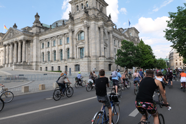 Eine Gruppe von Menschen, die auf Fahrrädern eine Straße vor dem Reichstaggebäude in Berlin, Deutschland, entlangfahren. Das Gebäude ist mit Fenstern, Säulen, Statuen und Flaggen geschmückt und von Bäumen und einem Geländer umgeben. Der Himmel ist bewölkt.