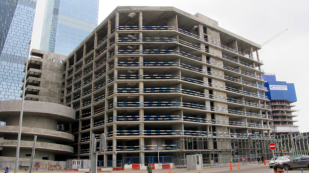 Ein großes Bauwerk im Bau in einer städtischen Umgebung, umgeben von Wolkenkratzern, Fahrzeugen, Fußgängern und Baumaschinen unter einem klaren blauen Himmel.
